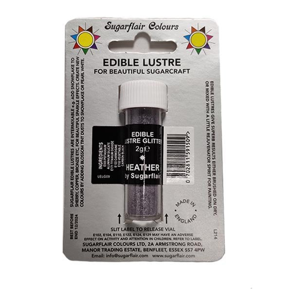 The image shows a small vial of edible lustre glitter labeled "Sugarflair Edible Lustre Glitter 2g Heather." The vial has a white screw cap and is prominently displayed on a card with colorful designs. The card features text that reads "EDIBLE LUSTRE FOR BEAUTIFUL SUGARCRAFT," along with details about the product, including the weight (2g) and its name "HEATHER." There are also ingredient and usage instructions, and it indicates that the product is made in England.