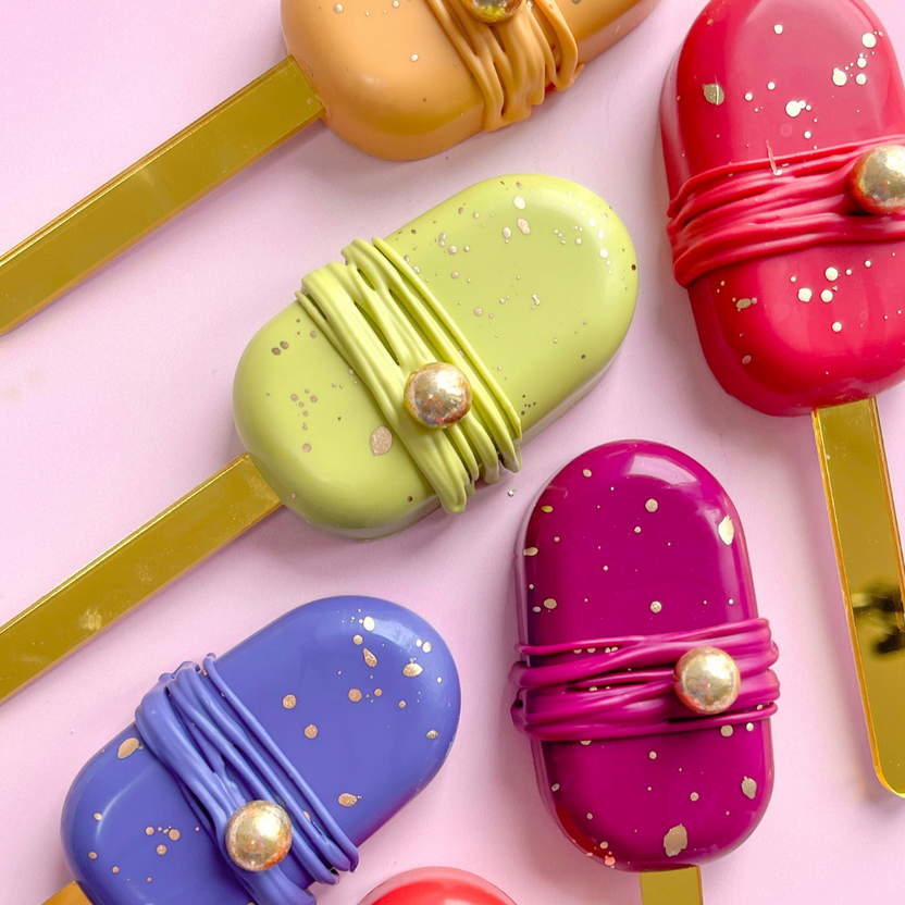 A flat lay of colorful popsicle-shaped treats arranged on a pink surface. The popsicles are in various colors, including green, orange, purple, red, and pink, with decorative elements like golden specks and ribbon-like adornments. Each popsicle has a light yellow stick at the bottom.