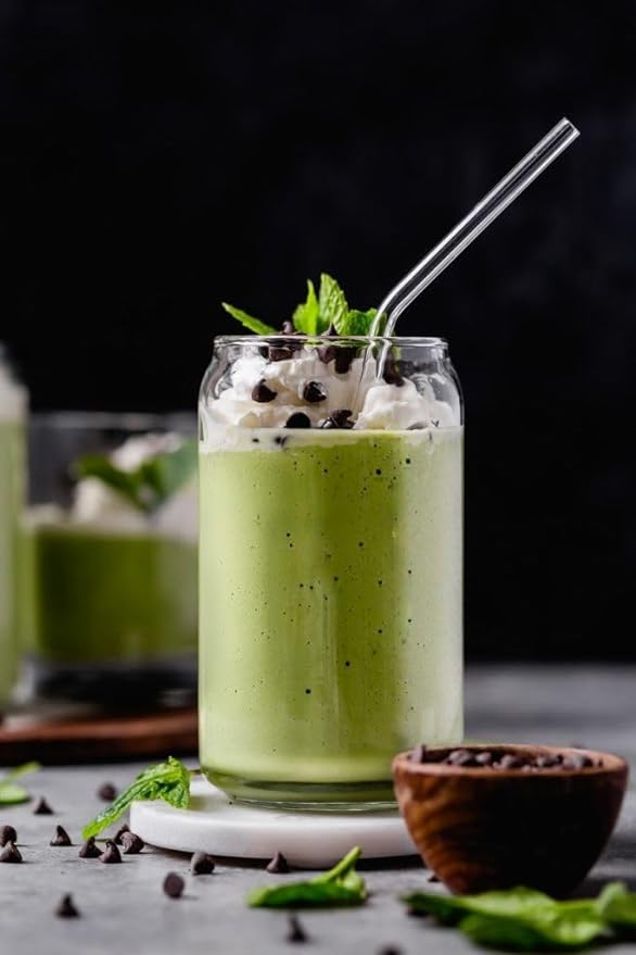 A glass jar filled with a creamy green beverage topped with whipped cream, mint leaves, and small chocolate chips. There is a clear straw in the drink. In the background, a similar glass is partially visible. The scene includes a small wooden bowl with chocolate chips and scattered mint leaves on a light-colored surface, creating a clean and elegant presentation.