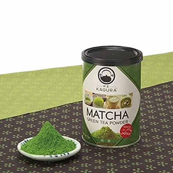A tin of Kagura Matcha Green Tea Powder (70g) is displayed with a small mound of green tea powder in a white dish beside it. The tin features a green label with the words "MATCHA GREEN TEA POWDER" and images of matcha drinks. The background is a patterned surface in dark brown, contrasting with a solid green tabletop.