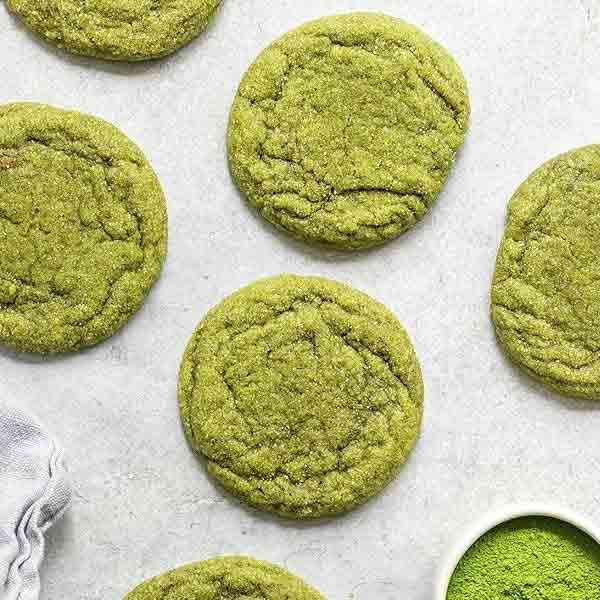 A flat lay image featuring several round, green cookies arranged on a light surface. The cookies have a textured surface and are made with matcha green tea. In the bottom right corner, there is a small white bowl filled with powdered green matcha tea. A gray cloth is partially visible on the left side of the image.