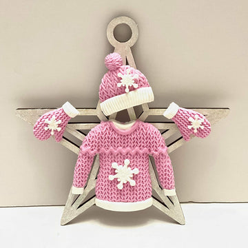 A decorative display featuring a pink winter sweater with a snowflake design, a matching pink hat, and pink mittens, all arranged on a white star-shaped background. The sweater features a textured knit pattern and the accessories are adorned with white details.