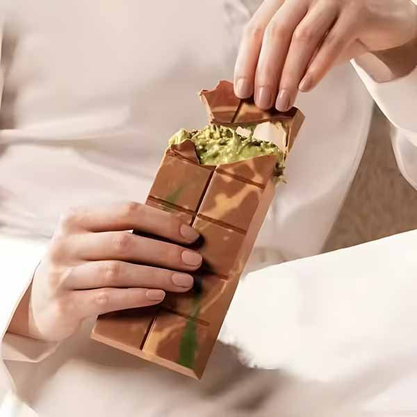 A person is holding a chocolate-shaped polycarbonate mold designed for making kunafa. The mold resembles a partially unwrapped chocolate bar, with a creamy green filling that appears to be the kunafa mixture visible inside. The person&