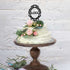 A three-tiered cake with a light green frosting is displayed on a wooden cake stand. The cake is adorned with pink roses, green leaves, and small berries. At the top of the cake, there is a black acrylic cake topper that features the word "LOVE" within an ornate frame. The background consists of a textured white wall.