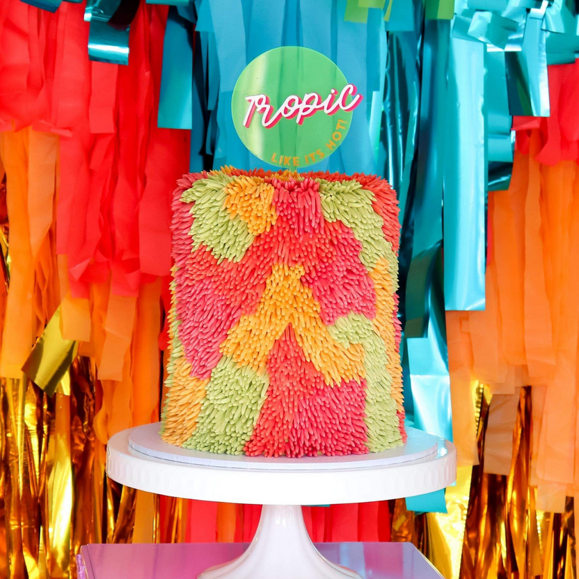 A colorful cake stands on a white pedestal, adorned with vibrant frosting in shades of pink, orange, and green, creating a textured floral appearance. Behind the cake, there is a festive backdrop of streamers in red, orange, blue, and gold. Prominently displayed on top of the cake is a circular sign that reads "Tropic" in pink lettering, accompanied by the phrase "LIKE IT&