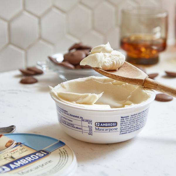 Open container of Ambrosi Mascarpone cheese with a wooden spoon scooping some out. The background features chocolate pieces, a glass with liquid, and a tiled wall, creating a cozy kitchen setting.