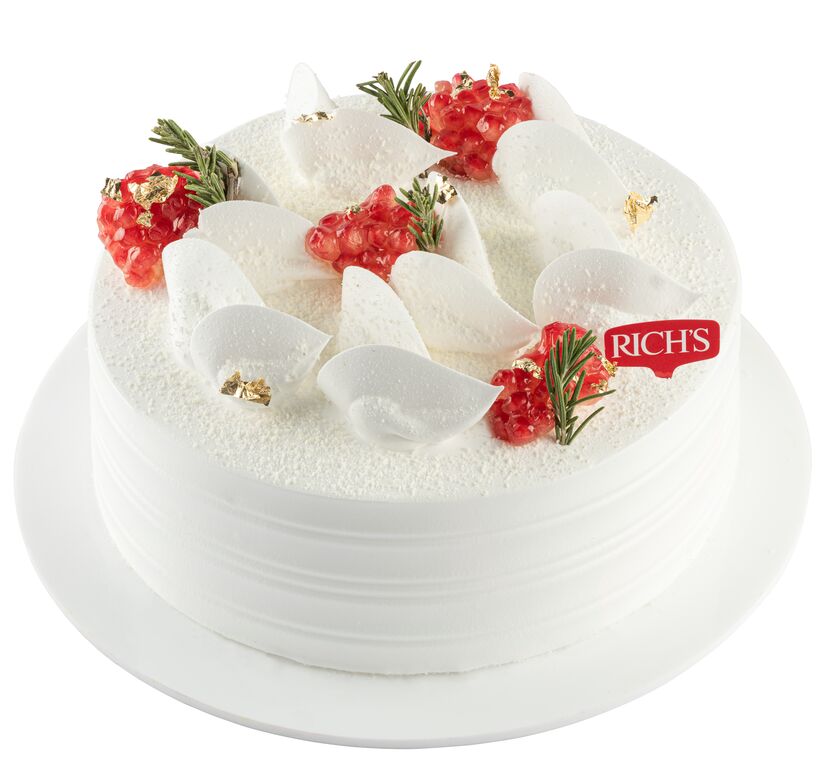 A round, white cake decorated with a smooth white frosting. The top is adorned with decorative elements including white chocolate or cream petals, small clusters of red jelly or fruit, and sprigs of green, possibly rosemary. There are accents of gold, adding a touch of elegance. A red &