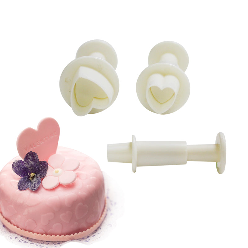 A set of three heart-shaped plunger cutters in white, displayed alongside a pink cake decorated with a heart-shaped topper that says "Valentine" and edible flowers. The cutters consist of two heart shapes and a plunger mechanism, showcasing their use for creating heart-shaped decorations. The cake features a textured surface with small heart designs.