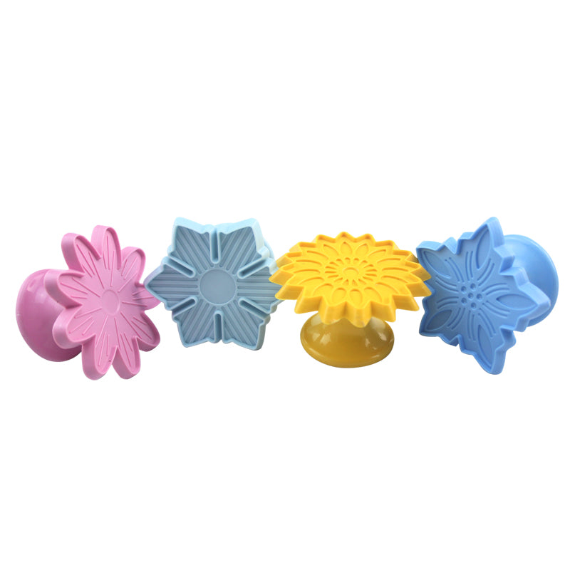 The image features a set of four plunger cutters, each designed to create solid shapes, specifically floral designs. The cutters vary in color: one is pink in the shape of a flower, a light blue one with a star-like shape, a yellow flower-shaped cutter, and another light blue cutter resembling a different flower. Each cutter has a raised design on top for imprinting patterns on dough or other materials, and they all have a sturdy base for easy handling.