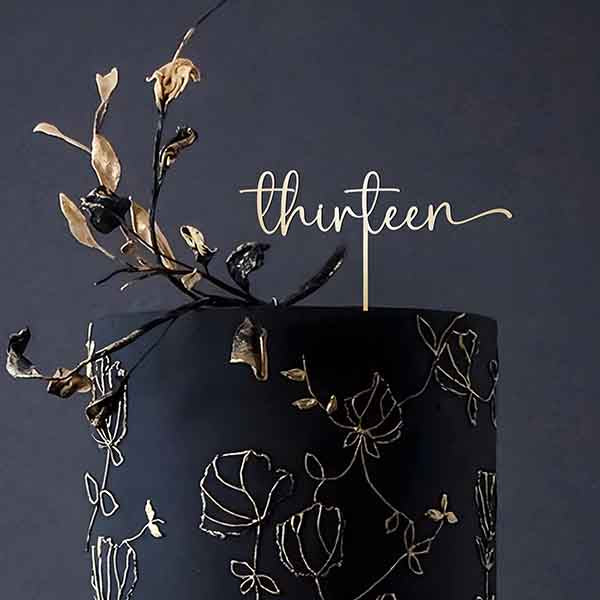 An elegant black cake decorated with floral designs. Atop the cake is a custom acrylic topper featuring the word "thirteen" in an elegant cursive font. The topper is complemented by delicate, artistic black and gold floral accents that extend from the cake&