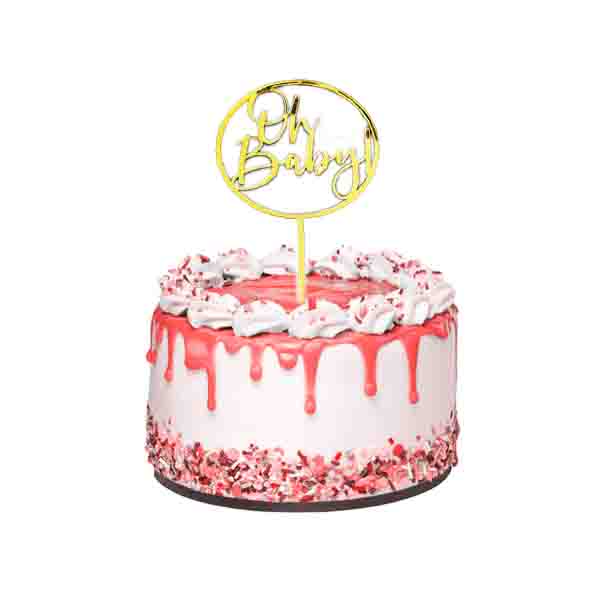 A decorated cake featuring a shiny gold acrylic topper that reads "Oh Baby!" The cake has a white base with pink icing drips and is topped with whipped cream and sprinkles. The sides of the cake are adorned with colorful sprinkles, creating a festive appearance.