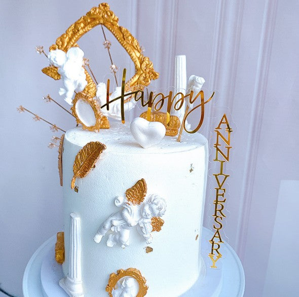 A beautifully decorated white cake featuring a variety of ornate accents. The cake is topped with an acrylic "Happy" and "ANNIVERSARY" topper in a golden script. Surrounding the toppers are decorative elements, including a golden frame, small white cherubs, a heart, and golden leaves. The cake has a smooth white frosting with scattered golden details, giving it an elegant and festive appearance. The overall design suggests a celebration, perfect for anniversaries.