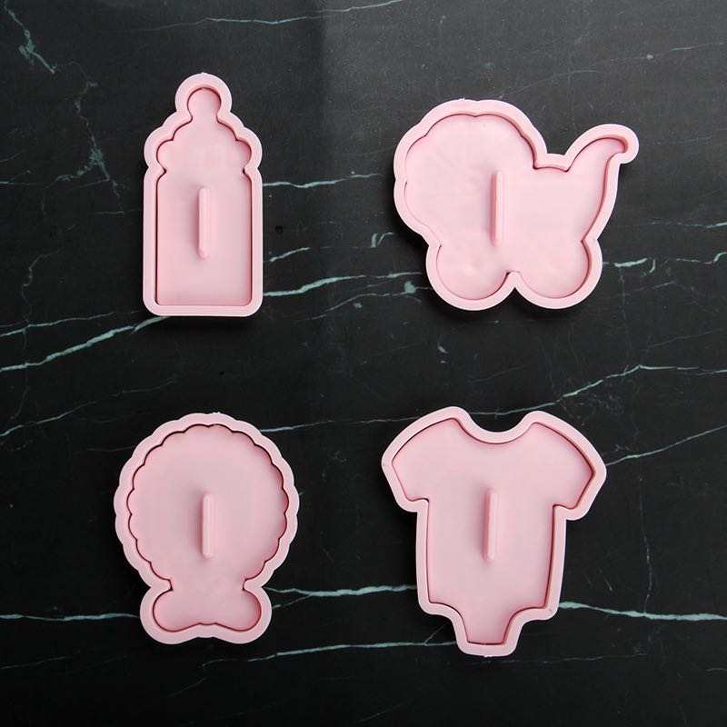 Four pink silicone cutters are arranged on a dark marble surface. The shapes include:
1. A baby bottle.
2. A stroller.
3. A round object resembling a gift or a decorative item.
4. A baby onesie.
Each cutter features a raised handle for ease of use.