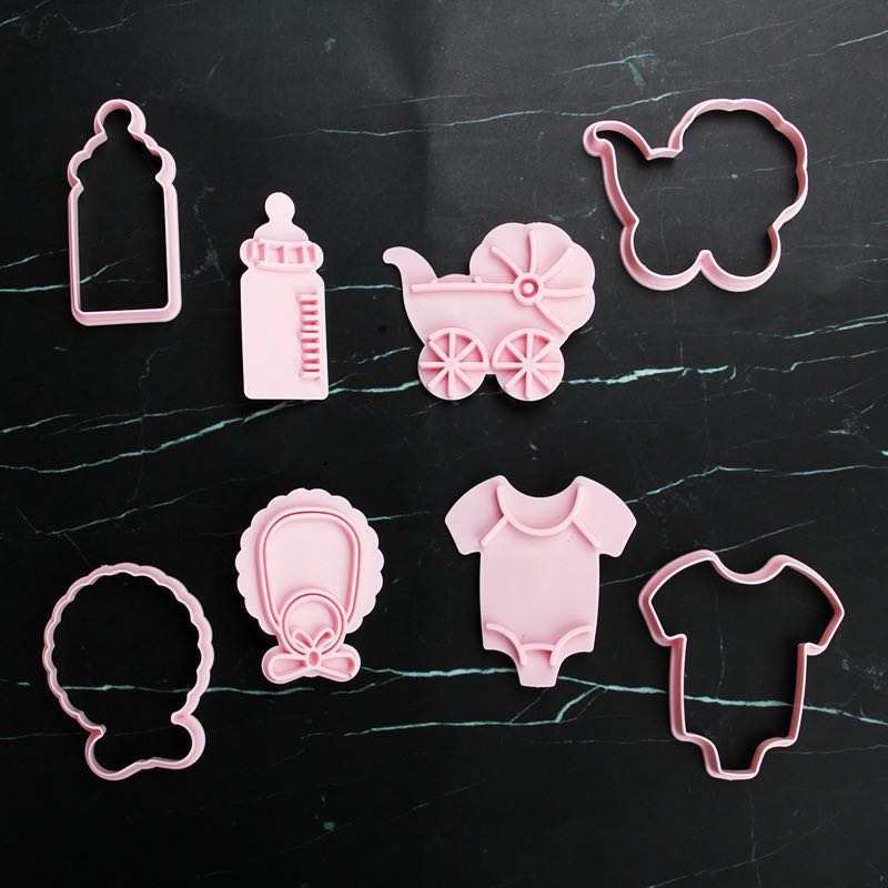 A collection of pink silicone cookie cutters arranged on a dark surface. The shapes include a baby bottle, a baby carriage, a bib, a onesie, and a butterfly. The cutters vary in design and size, all featuring a soft pink color.