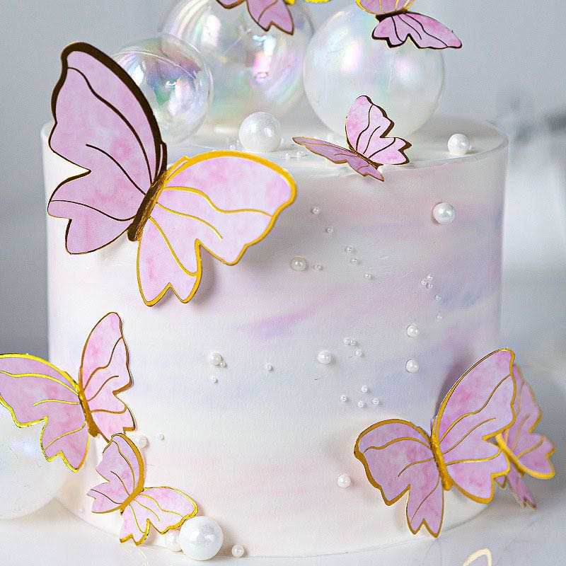A decorative cake features a smooth, white surface with a subtle iridescent sheen. Multiple pink butterfly toppers, outlined in gold, are artfully arranged on the cake, some positioned on the sides and others on the top. The butterflies vary in size and orientation. Additionally, the cake is adorned with shiny, clear bubbles and small, round, white decorations that resemble pearls, enhancing its whimsical look.