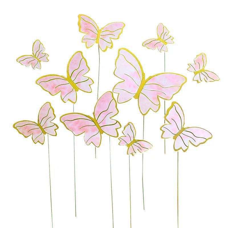A collection of decorative butterfly cake toppers in various sizes. Each butterfly features a delicate design with soft pink wings and gold outlines. The butterflies are attached to thin white sticks for easy placement on cakes or desserts, arranged in an expressive, scattered formation.