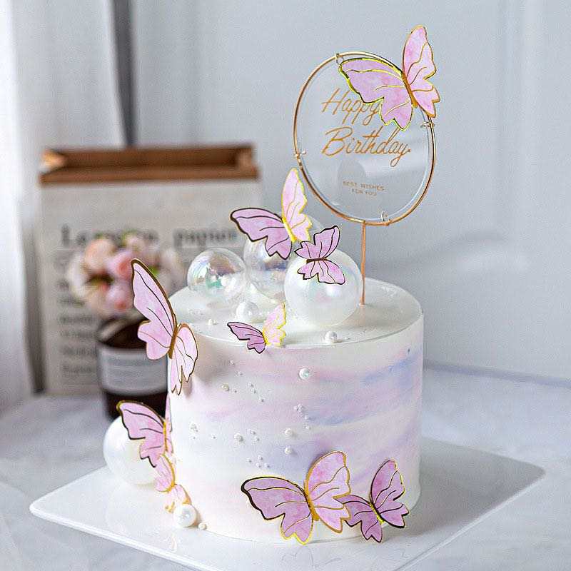A beautifully decorated cake with a smooth white surface adorned with colorful butterfly cake toppers in pink and gold. The cake features a round topper that reads "Happy Birthday" surrounded by several decorative bubbles and a few additional pearls. The background includes a blurred flower arrangement and a piece of wall art, suggesting a celebratory setting.