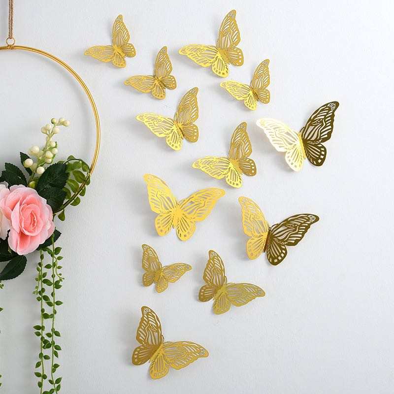 A collection of decorative butterfly stickers in various sizes and designs, primarily in gold, with a few white and brown accents. The butterflies are arranged on a light-colored wall, with a floral arrangement featuring pink roses and greenery in the foreground, partially visible.