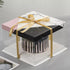 A tall cake box with a square base and a marble-patterned lid is shown. The box features a combination of black, white, and pink colors, with a satin gold ribbon tied around it. Inside, there is a cake with a decorative dripping design. The box is set on a light-colored surface, with some blurred items in the background.