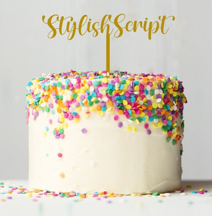A round cake with white frosting, topped with colorful sprinkles in shades of pink, blue, yellow, and green. Atop the cake, there is a golden cake topper that reads "Stylish Script," standing upright on a thin golden stick. The background is plain, emphasizing the cake and its decorations.