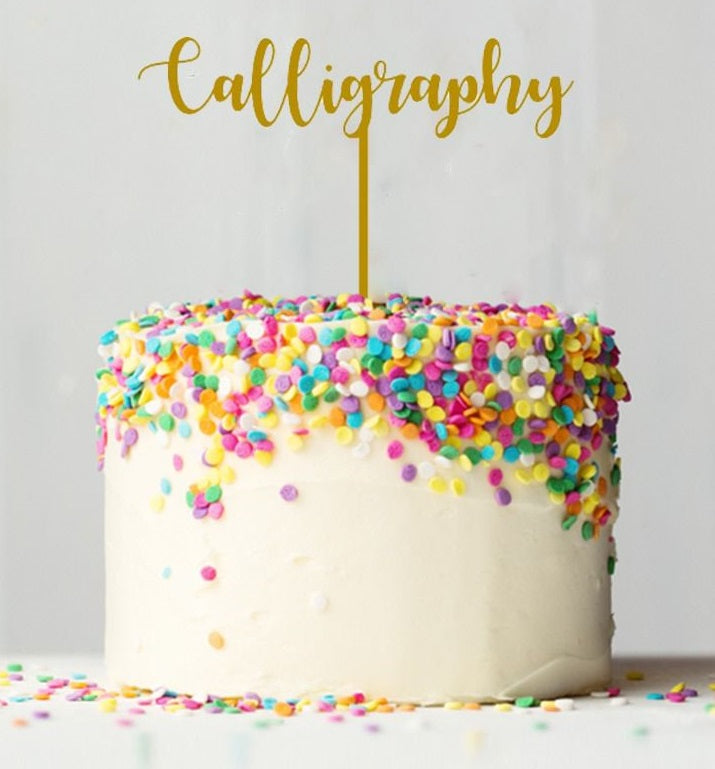 A round, white cake topped with colorful sprinkles in shades of pink, blue, yellow, and green. Atop the cake is a gold card stock topper that reads "Calligraphy" in an elegant font. The background is light and neutral, enhancing the colorful decorations on the cake.