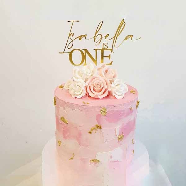 A pink cake adorned with a gold acrylic topper that reads "Isabella is ONE," featuring decorative roses in white and soft pink on top. The cake is decorated with a textured finish and subtle gold accents, all set against a light background.