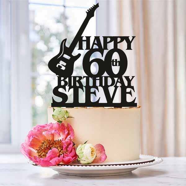 A birthday cake decorated with a black acrylic cake topper featuring a guitar design and the words "HAPPY 60th BIRTHDAY STEVE." The cake is adorned with pink and cream-colored flowers at its base and is presented on a white plate. The background shows a bright and softly lit interior setting.