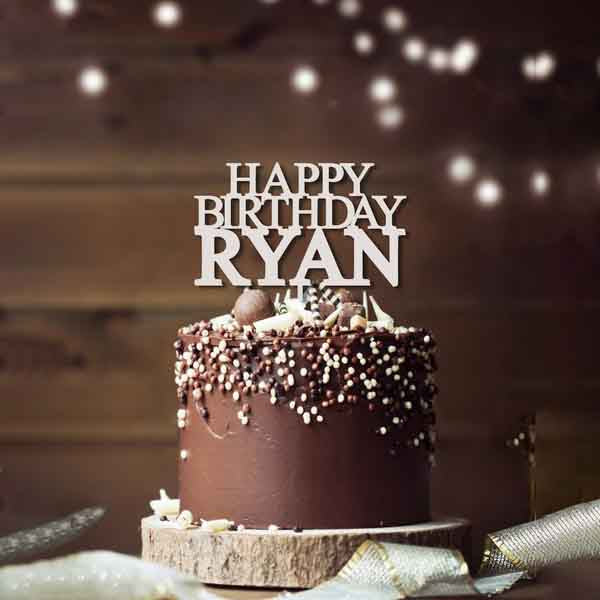 A chocolate birthday cake topped with a personalized acrylic cake topper that reads "HAPPY BIRTHDAY RYAN." The cake is decorated with chocolate sprinkles and nuts, resting on a wooden base. Soft bokeh lights are visible in the background, creating a festive atmosphere.