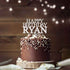 A chocolate birthday cake topped with a personalized acrylic cake topper that reads "HAPPY BIRTHDAY RYAN." The cake is decorated with chocolate sprinkles and nuts, resting on a wooden base. Soft bokeh lights are visible in the background, creating a festive atmosphere.