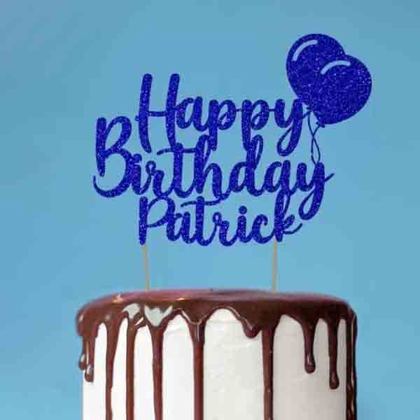 A birthday cake with a glossy chocolate drip on the edges. Topped with a glittery blue cake topper that reads "Happy Birthday Patrick," accompanied by a blue balloon graphic, against a light blue background.