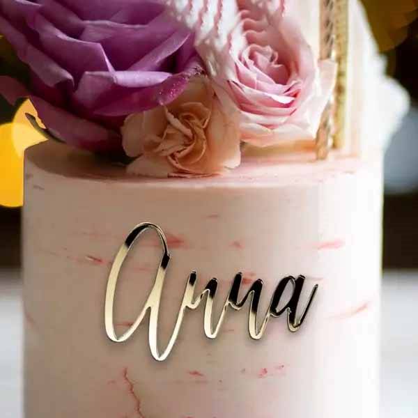 An elegant cake is displayed, featuring a smooth pink frosting with subtle marbling. At the base of the cake sits a personalized name charm topper reading "Anna" in a stylish, cursive font. The cake is adorned with pastel-colored flowers, including purple and pink roses, adding a decorative touch. The background is softly blurred, emphasizing the cake and its decorative elements.