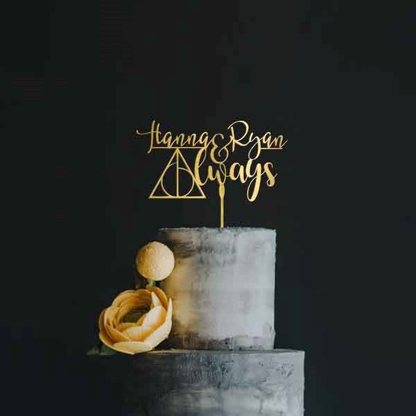 A stylish wedding cake featuring a gold acrylic cake topper that reads "Hanna & Ryan Always," with a decorative triangle symbol in the center. The cake has a smooth, gray finish and is embellished with a soft yellow rose and a small round decorative element on the side. The background is a simple dark color, enhancing the contrast of the cake and topper.