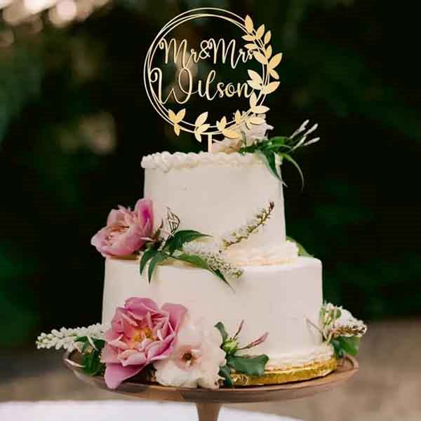 An elegant three-tiered white wedding cake is adorned with a gold acrylic cake topper that reads "Mr & Mr Wilson" surrounded by decorative leaves. The cake is decorated with fresh pink and white flowers and greenery, elegantly placed on a round cake stand. The background features a soft, natural setting, enhancing the romantic atmosphere of the display.