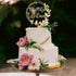 An elegant three-tiered white wedding cake is adorned with a gold acrylic cake topper that reads "Mr & Mr Wilson" surrounded by decorative leaves. The cake is decorated with fresh pink and white flowers and greenery, elegantly placed on a round cake stand. The background features a soft, natural setting, enhancing the romantic atmosphere of the display.