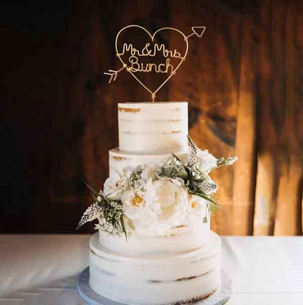 A three-tiered white wedding cake with a rustic finish features a personalized acrylic cake topper. The topper is shaped like a heart and displays the text "Mr & Mrs Bunch" in an elegant script. The cake is adorned with white and yellow flowers, along with greenery, and sits on a silver cake board. The background shows a warm wood paneling, enhancing the romantic ambiance.