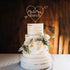 A three-tiered white wedding cake with a rustic finish features a personalized acrylic cake topper. The topper is shaped like a heart and displays the text "Mr & Mrs Bunch" in an elegant script. The cake is adorned with white and yellow flowers, along with greenery, and sits on a silver cake board. The background shows a warm wood paneling, enhancing the romantic ambiance.