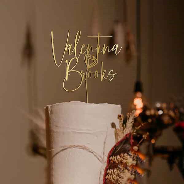 A close-up of a wedding cake with a personalized cake topper. The topper features the name "Valentina Brooks" in an elegant, cursive gold font, accompanied by a simple flower illustration. The cake appears white with a textured finish, and there are decorative elements, including dried flowers, surrounding the base. The background is softly blurred with warm tones and hanging lights, creating a romantic atmosphere.