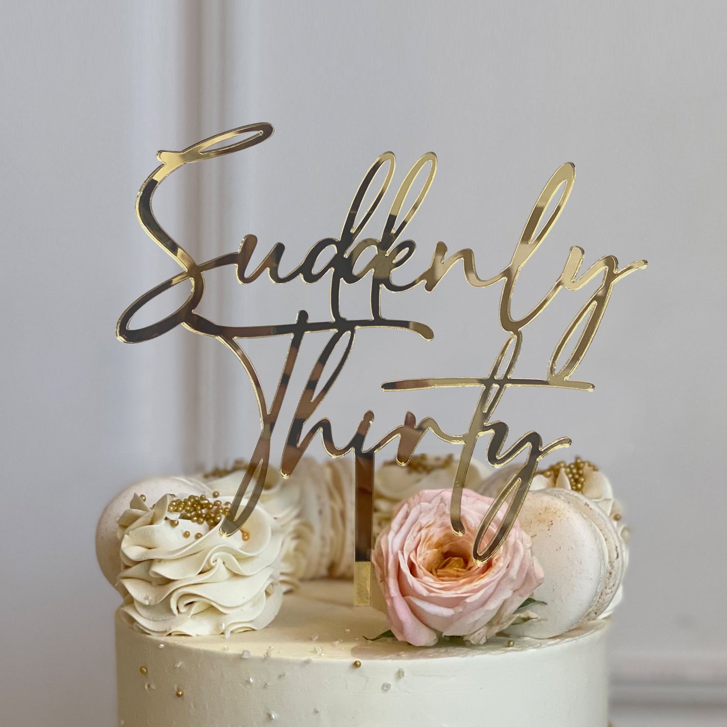 An elegant cake with a smooth, light-colored frosting is topped with a customized acrylic cake topper that reads "Suddenly Thirty" in a stylish, cursive font. The topper is golden and stands prominently above the cake. Surrounding the topper, the cake features decorative elements such as swirls of cream, edible gold pearls, a pink rose, and white macarons. The overall presentation suggests a celebratory theme, suitable for a 30th birthday party.