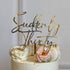 An elegant cake with a smooth, light-colored frosting is topped with a customized acrylic cake topper that reads "Suddenly Thirty" in a stylish, cursive font. The topper is golden and stands prominently above the cake. Surrounding the topper, the cake features decorative elements such as swirls of cream, edible gold pearls, a pink rose, and white macarons. The overall presentation suggests a celebratory theme, suitable for a 30th birthday party.