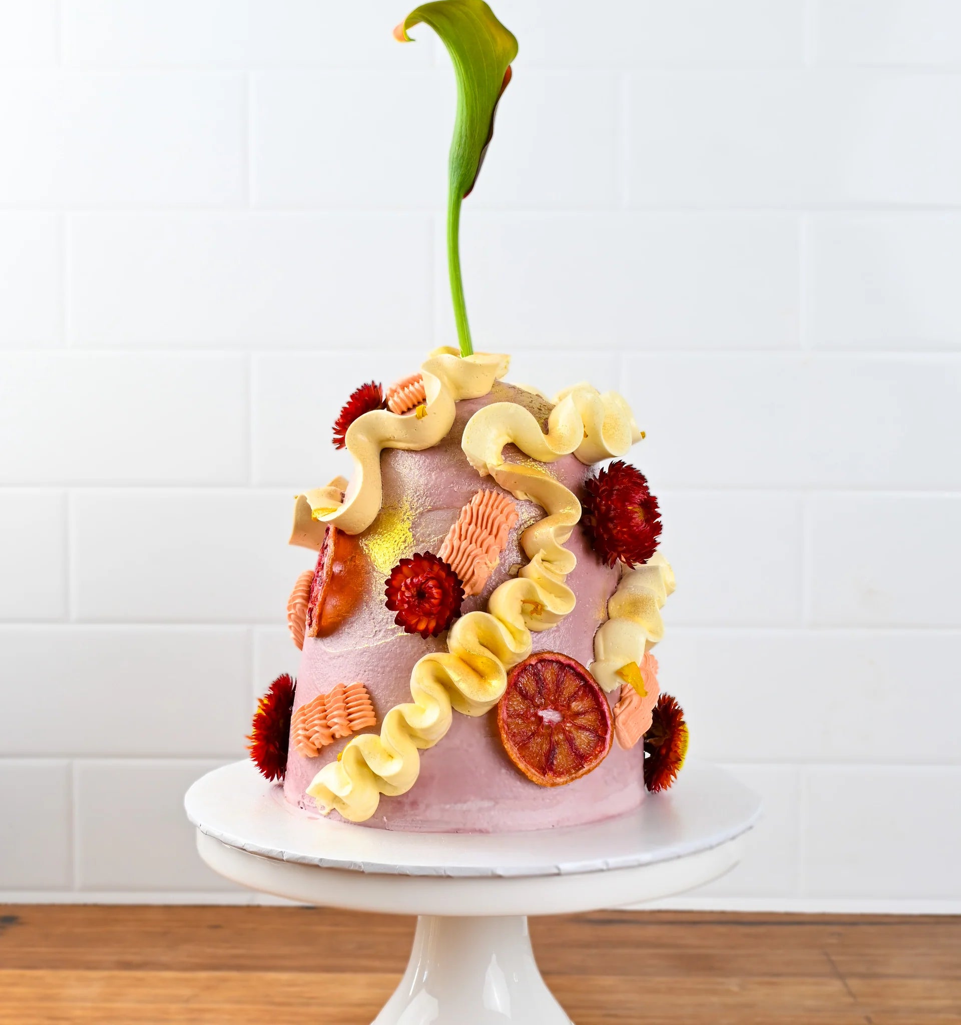 A beautifully decorated cake on a white pedestal. The cake is pink with a smooth finish and adorned with various toppings, including yellow and orange ruffled decorations, bright red edible flowers, and slices of dried fruit. At the top, a tall green plant stem adds an artistic touch. The background features white tiled walls.
