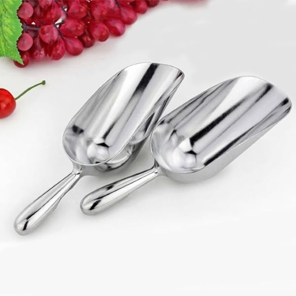 A pair of stainless steel scoops are displayed on a light background, with one scoop slightly overlapping the other. The scoops have polished, shiny surfaces and curved bowls, suitable for scooping flour or other ingredients. In the background, there are red grapes and green leaves, adding a natural touch to the scene.