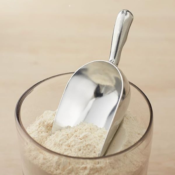 A stainless steel scoop is resting on top of a glass container filled with flour. The scoop has a smooth, shiny surface and a curved shape, partially submerged in the fine white powder. The background is a light wooden surface.