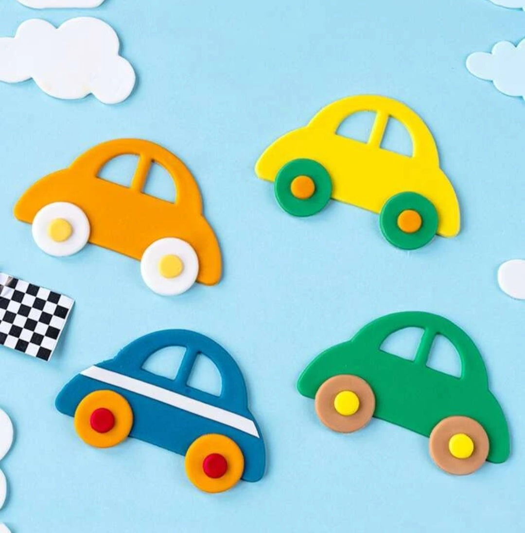 The image features several colorful cookie cutouts shaped like cars, placed on a light blue background. There are four car shapes in total: an orange car with white wheels, a yellow car with green wheels, a blue car with a white stripe and red wheels, and a green car with brown wheels. Additionally, there are a few white cloud shapes scattered around, and a black and white checkered flag in the corner.