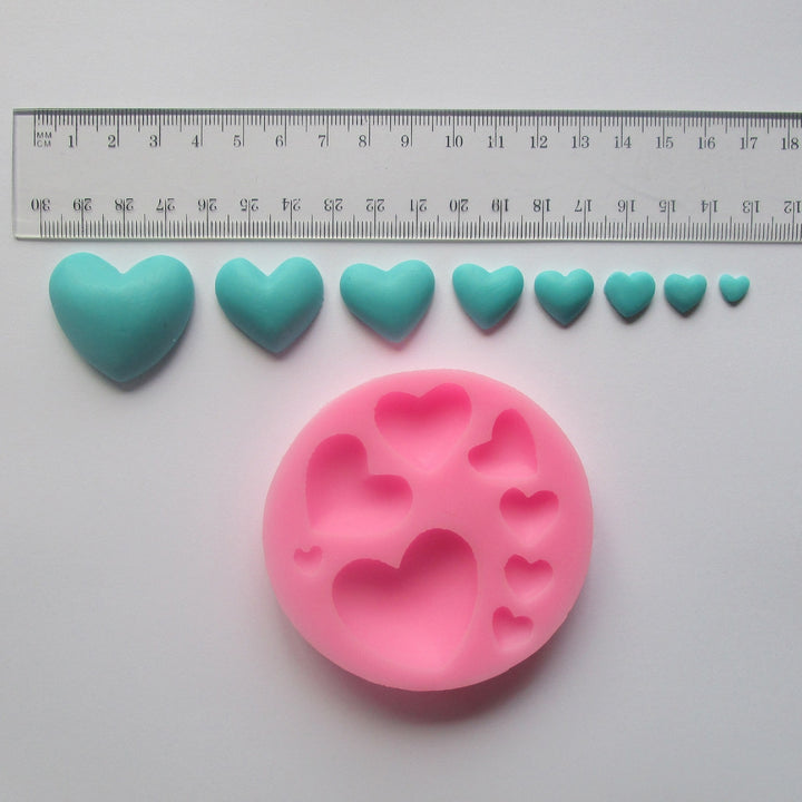 A pink silicone mould shaped like hearts is displayed on a flat surface, with several small turquoise heart-shaped pieces made from a similar material arranged above it. A ruler is placed above the hearts for scale, showing measurements in centimeters and inches. The silicone mould has various sizes of heart shapes indented into its surface.