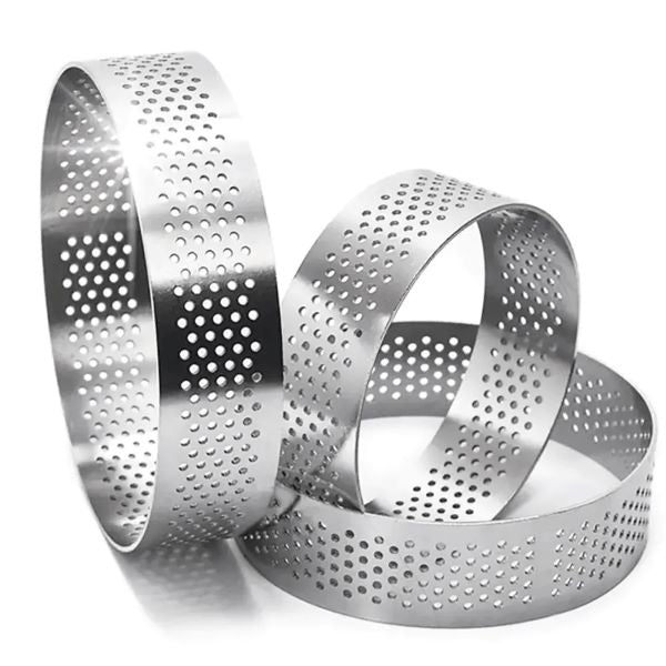 Three stainless steel perforated tart rings of varying heights and sizes. Each ring has a shiny metallic finish and features evenly spaced circular holes throughout its surface. The rings are stacked and placed at different angles, showcasing their round shape and design. The dimensions of the ring in focus are approximately 9 cm in diameter and 2.5 cm in height.