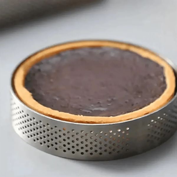 A stainless steel perforated tart ring sitting on a flat surface, holding a tart with a dark chocolate filling and a golden-brown crust. The tart ring has a diameter of 9 cm and a height of 2.5 cm, with small holes around its sides. The background is neutral and slightly blurred.