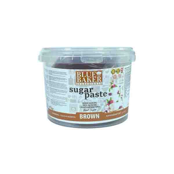 A transparent plastic container of "Blue Baker Brown Sugarpaste Fondant." The container features a white label with the text "sugar paste" prominently displayed, along with the brand name "Blue Baker" at the top. The label also indicates the color of the product as "BROWN." Additional information, likely including ingredients and usage instructions, is printed on the sides of the container. The contents appear dark brown, suggesting a rich color typical of brown sugarpaste.