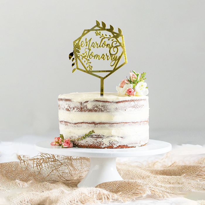 A layered naked cake is displayed on a white cake stand. The cake has visible layers with cream frosting between them. A gold acrylic cake topper, shaped like a hexagon, sits atop the cake and features the names "Marlon & Tomarry" in an elegant script. Delicate flowers, including small pink roses and white blooms, are arranged on the cake next to the topper, adding a decorative touch. The background is softly blurred, emphasizing the cake.