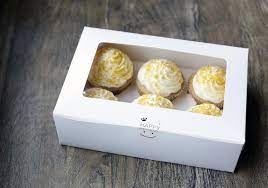 A white cardboard cupcake box with a clear window, containing six cupcakes with creamy yellow frosting, placed on a wooden surface. The box is designed to hold six portions and features a cheerful, minimalistic design.