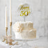 A two-tiered white cake decorated with smooth, swirled icing sits on a clear cake stand. Atop the cake is a circular cake topper that reads "Happy 50th" in elegant gold lettering. A large, pale rose is positioned on the side of the cake. In the background, there are two tall white candles and an ornate, vintage-style holder. The setting features a soft, neutral backdrop to highlight the cake and decorations.